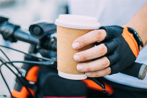 A hand holding a paper cup with hot beverage with one hand and the handlebar of a bike with the other